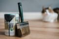 Professional paint brush near metal can with information and dirty flows on parquet in house